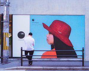 A person wearing a white shirt and black cap stands with their back to the camera in front of a large advertisement. The advertisement features a profile of a woman wearing a wide-brimmed red hat and an orange top against a bright blue background. The setting appears to be an urban area with a sidewalk and a metal railing.