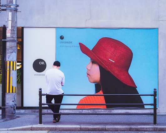A person wearing a white shirt and black cap stands with their back to the camera in front of a large advertisement. The advertisement features a profile of a woman wearing a wide-brimmed red hat and an orange top against a bright blue background. The setting appears to be an urban area with a sidewalk and a metal railing.