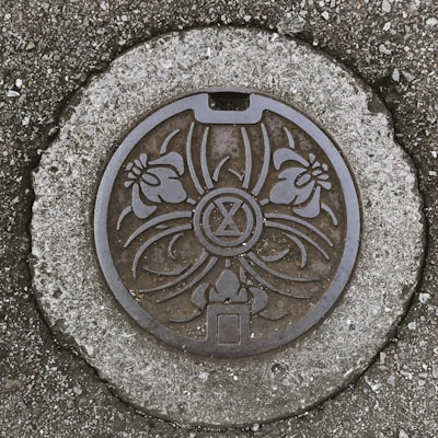 A round manhole cover with an intricate design featuring floral patterns and an hourglass-like symbol in the center. The cover is set in textured concrete surrounded by coarse, small stones.
