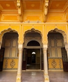 An ornate architectural space with intricately designed archways and pillars featuring yellow and gold tones. The ceiling and walls display exquisite geometric and floral patterns, adding to the richness of the ambiance. There are wooden doors leading into interior rooms, creating a sense of depth and mystery.