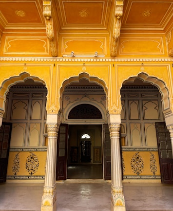 An ornate architectural space with intricately designed archways and pillars featuring yellow and gold tones. The ceiling and walls display exquisite geometric and floral patterns, adding to the richness of the ambiance. There are wooden doors leading into interior rooms, creating a sense of depth and mystery.