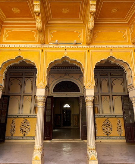 An ornate architectural space with intricately designed archways and pillars featuring yellow and gold tones. The ceiling and walls display exquisite geometric and floral patterns, adding to the richness of the ambiance. There are wooden doors leading into interior rooms, creating a sense of depth and mystery.