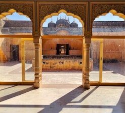 A traditional architectural setting featuring arched windows with intricate carvings and a view of an open courtyard. The foreground includes ornately decorated columns, while through the arches, a rustic, weathered wall and structure are visible.