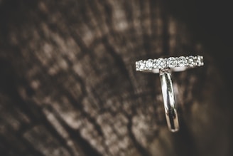 A silver-colored ring adorned with multiple small diamonds resting on a dark and textured wood surface, creating a contrast between the sparkling jewels and the natural background.