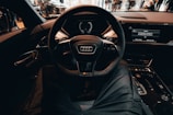 A view from the driver's seat of a modern car, focusing on the steering wheel with an Audi logo. The dashboard displays digital screens, and the interior appears sleek and high-tech. People are visible outside through the car windows, indicating a public or exhibition setting.