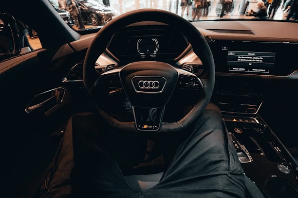 A view from the driver's seat of a modern car, focusing on the steering wheel with an Audi logo. The dashboard displays digital screens, and the interior appears sleek and high-tech. People are visible outside through the car windows, indicating a public or exhibition setting.