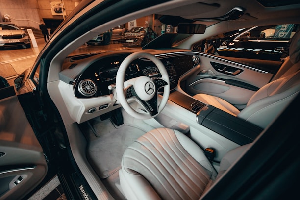 A luxurious car interior featuring high-end leather seats, a sleek dashboard with digital displays, and a stylish steering wheel with the Mercedes-Benz logo. The design is modern and sophisticated, with ambient lighting adding to the premium feel.