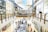 A bustling shopping mall with two levels, adorned with festive decorations. Shoppers are browsing various stores and kiosks, with a Christmas tree visible among the stalls. The upper level has glass railings and shops lining both sides, including well-known brands.