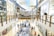 A bustling shopping mall with two levels, adorned with festive decorations. Shoppers are browsing various stores and kiosks, with a Christmas tree visible among the stalls. The upper level has glass railings and shops lining both sides, including well-known brands.