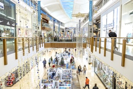 A bustling shopping mall with two levels, adorned with festive decorations. Shoppers are browsing various stores and kiosks, with a Christmas tree visible among the stalls. The upper level has glass railings and shops lining both sides, including well-known brands.