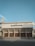 An auditorium building with a flat roof and large wooden doors at the entrance. The structure has a modern architectural style, with prominent vertical columns. A person is walking by on the pavement in front of the building. The sky is clear with a few clouds visible.