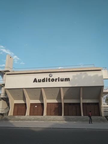 An auditorium building with a flat roof and large wooden doors at the entrance. The structure has a modern architectural style, with prominent vertical columns. A person is walking by on the pavement in front of the building. The sky is clear with a few clouds visible.