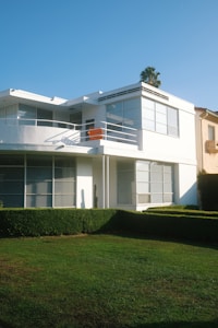 A modern, two-story house with large windows and a flat roof. The facade is primarily white, and there is a small balcony with white railings and an orange chair. The foreground features a well-manicured lawn and neatly trimmed hedges, while a palm tree is visible behind the house.