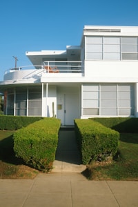 A modernist-style two-story building with large windows and a flat roof. The building is white with sharp, clean lines and features a balcony on the upper floor. Neatly trimmed hedges line the pathway leading to the front door, enhancing the minimalist and organized aesthetic.