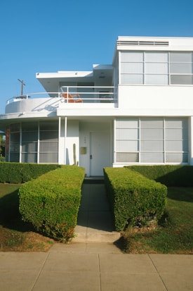A modernist-style two-story building with large windows and a flat roof. The building is white with sharp, clean lines and features a balcony on the upper floor. Neatly trimmed hedges line the pathway leading to the front door, enhancing the minimalist and organized aesthetic.