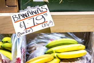 A display shelf holds a bunch of bananas, with a price tag indicating £1.79 per kilogram from the Dominican Republic. The bananas are in plastic bags, with some still green and others ripe yellow.