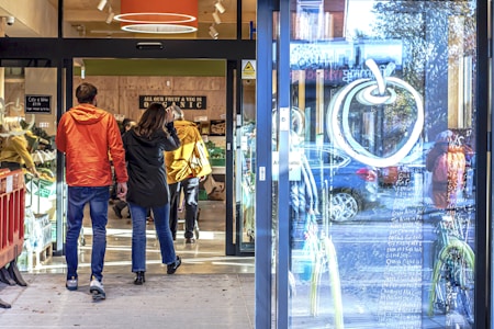 A couple walks into an organic grocery store. The entrance is glass with a large apple design and handwritten notes visible on the door. The interior shows various organic produce and a sign indicating organic goods. Natural light and reflections are apparent on the glass.