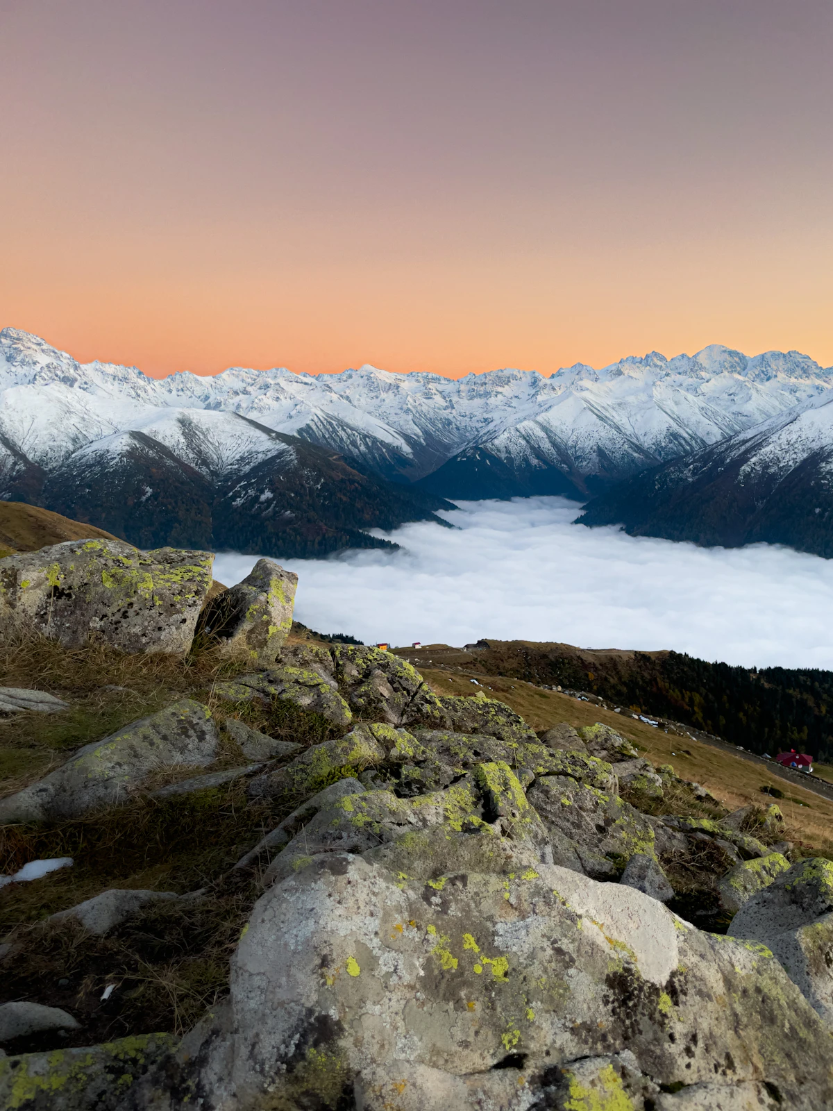 Snow-covered peaks of the Kaçkar range under an orange sky, above a sea of cloud, seen from lichen-covered rocks at Huser