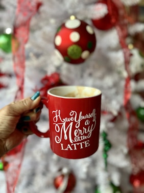 A hand holds a red mug with white script text that reads 'Have Yourself a Merry little LATTE' with an out-of-focus Christmas setting in the background, featuring a white tree adorned with colorful ornaments.