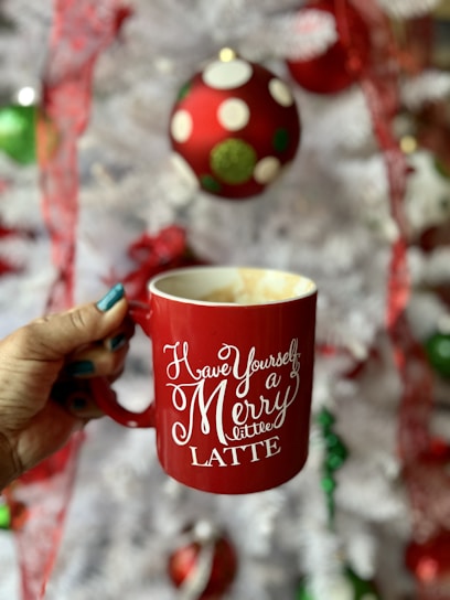 A hand holds a red mug with white script text that reads 'Have Yourself a Merry little LATTE' with an out-of-focus Christmas setting in the background, featuring a white tree adorned with colorful ornaments.