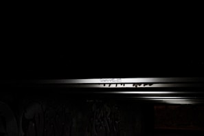 A dimly lit urban setting with graffiti on a concrete wall. The image is predominantly dark with a light source illuminating a section of the wall, revealing spray-painted artwork and scribbled text. The beams of light create a contrast between the illuminated area and the surrounding darkness.