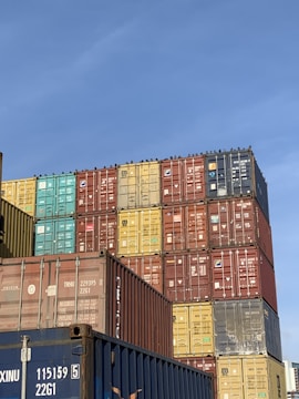 A large stack of colorful shipping containers arranged in an organized manner under a clear blue sky. The containers feature various colors including red, yellow, green, blue, and brown, with some brand logos and identification numbers visible.