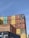 A large stack of colorful shipping containers arranged in an organized manner under a clear blue sky. The containers feature various colors including red, yellow, green, blue, and brown, with some brand logos and identification numbers visible.