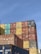 A large stack of colorful shipping containers arranged in an organized manner under a clear blue sky. The containers feature various colors including red, yellow, green, blue, and brown, with some brand logos and identification numbers visible.
