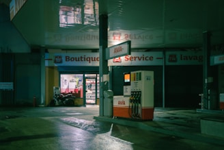 A dimly lit gas station at night with a visible convenience store labeled 'Boutique'. The station includes a set of fuel pumps with an illuminated Avia sign. The area is mostly empty except for a stationary motorcycle near the entrance of the store, which has its interior lights on, displaying products inside.