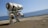 A futuristic robot with a bulky, mechanical design stands on a paved surface near a body of water under a clear blue sky. The robot is primarily white with blue accents and features a rounded body supported by two substantial legs. Various joints and mechanical parts are visible, indicative of advanced technology.