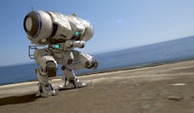 A futuristic robot with a bulky, mechanical design stands on a paved surface near a body of water under a clear blue sky. The robot is primarily white with blue accents and features a rounded body supported by two substantial legs. Various joints and mechanical parts are visible, indicative of advanced technology.