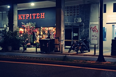 A flower shop with a bright sign in Greek, displaying a colorful array of flowers and plants outside. Two motorcycles are parked nearby under dim streetlights, with some graffiti visible on the building wall next to the shop.