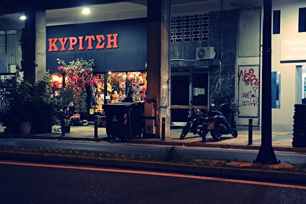 A flower shop with a bright sign in Greek, displaying a colorful array of flowers and plants outside. Two motorcycles are parked nearby under dim streetlights, with some graffiti visible on the building wall next to the shop.