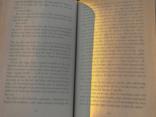 An open book lies flat, displaying pages 10 and 11. The text is well-lit, with a shadow and sunlight creating a contrast on the right side. The narrative appears to be about a cafe closing down, daily routines, and personal reflections.
