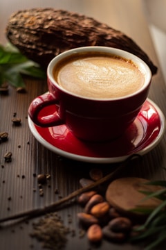 A red coffee cup sits on a saucer filled with steaming coffee. Surrounding the cup are cacao beans, a piece of cocoa pod, and some leaves, suggesting a coffee or chocolate theme. The setting is wooden, possibly a table, and has a rustic, cozy feel.