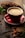 A red coffee cup sits on a saucer filled with steaming coffee. Surrounding the cup are cacao beans, a piece of cocoa pod, and some leaves, suggesting a coffee or chocolate theme. The setting is wooden, possibly a table, and has a rustic, cozy feel.