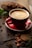 A red coffee cup sits on a saucer filled with steaming coffee. Surrounding the cup are cacao beans, a piece of cocoa pod, and some leaves, suggesting a coffee or chocolate theme. The setting is wooden, possibly a table, and has a rustic, cozy feel.