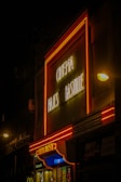 A vintage-looking cinema facade with bright neon lights prominently displaying 'Cinema Majestic Bastille' in a classic script. The building is illuminated with red and yellow neon, casting a warm glow onto the street. Below the main sign, movie posters and additional signage are visible, hinting at a lively urban environment.