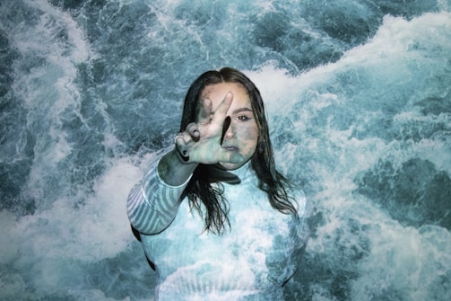 A person is standing in front of an image of turbulent water. The water is projected onto their body and face, creating a blending effect where the person almost merges with the background. The individual has their right hand raised in front of their face, with fingers partially spread as if blocking something or shielding themselves.