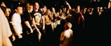 A large group of people, mostly young adults, are gathered in a lively environment that resembles a concert or a club. The individuals appear to be engaging with one another energetically, with one person kneeling in the middle as others stand around him. The scene is dimly lit, creating a warm and dynamic atmosphere.