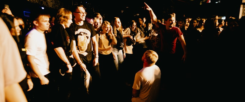 A large group of people, mostly young adults, are gathered in a lively environment that resembles a concert or a club. The individuals appear to be engaging with one another energetically, with one person kneeling in the middle as others stand around him. The scene is dimly lit, creating a warm and dynamic atmosphere.