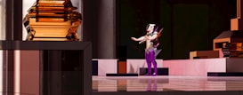 A futuristic scene with a humanoid figure in the center, wearing an elaborate costume with vibrant purple and white colors. The background and surrounding environment feature geometric shapes and metallic surfaces, with an amber-colored structure prominently on the left.