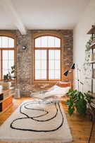 A stylish living room with large arched windows set in a brick wall. The room features a modern lounge chair with a sheepskin throw, a contemporary patterned rug, and various plants. A wooden floor and a mix of white and brick walls add to the cozy and inviting atmosphere.