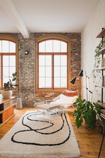 A stylish living room with large arched windows set in a brick wall. The room features a modern lounge chair with a sheepskin throw, a contemporary patterned rug, and various plants. A wooden floor and a mix of white and brick walls add to the cozy and inviting atmosphere.