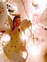 A handcrafted angel ornament with a heart design is placed on a brightly lit Christmas tree with white branches. The angel has a halo, painted features, and polka dot detailing.