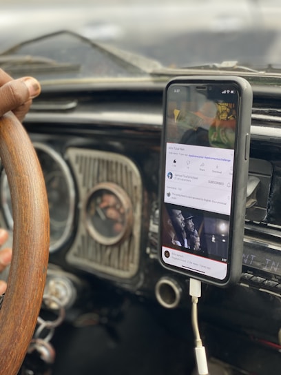 A person is holding a wooden steering wheel while a smartphone is mounted on the dashboard of a car. The phone is playing a YouTube video, and it is connected via a charging cable.