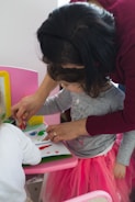 A young child engages in a creative activity, painting with their fingers on paper, while an adult assists and guides them. The child, adorned in a pink tutu-like skirt, appears focused on the task. The setting includes a pink chair and a bright yellow frame in the background.