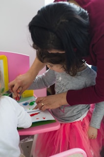 A young child engages in a creative activity, painting with their fingers on paper, while an adult assists and guides them. The child, adorned in a pink tutu-like skirt, appears focused on the task. The setting includes a pink chair and a bright yellow frame in the background.