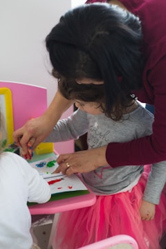 A young child engages in a creative activity, painting with their fingers on paper, while an adult assists and guides them. The child, adorned in a pink tutu-like skirt, appears focused on the task. The setting includes a pink chair and a bright yellow frame in the background.