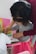 A young child engages in a creative activity, painting with their fingers on paper, while an adult assists and guides them. The child, adorned in a pink tutu-like skirt, appears focused on the task. The setting includes a pink chair and a bright yellow frame in the background.